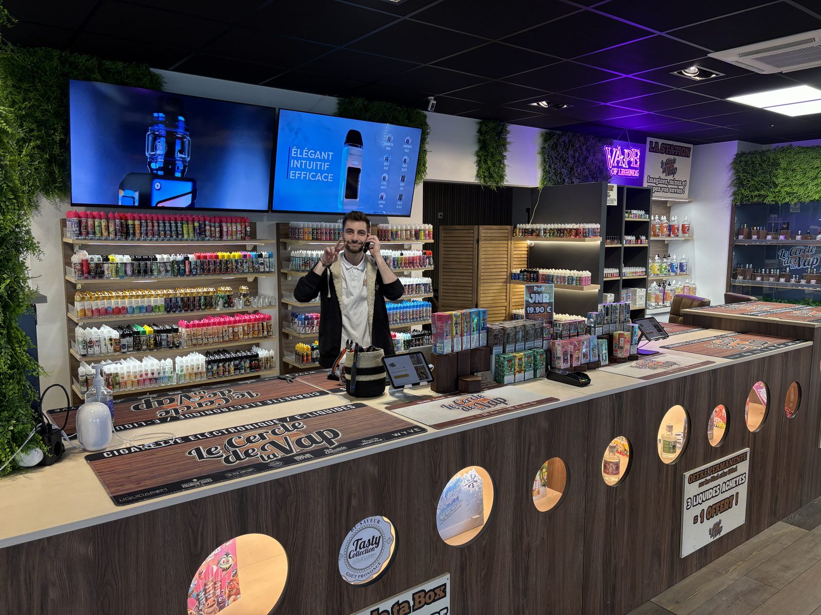 Comptoir de la boutique avec écrans de présentation, linéaires d'e-liquides et un conseiller souriant